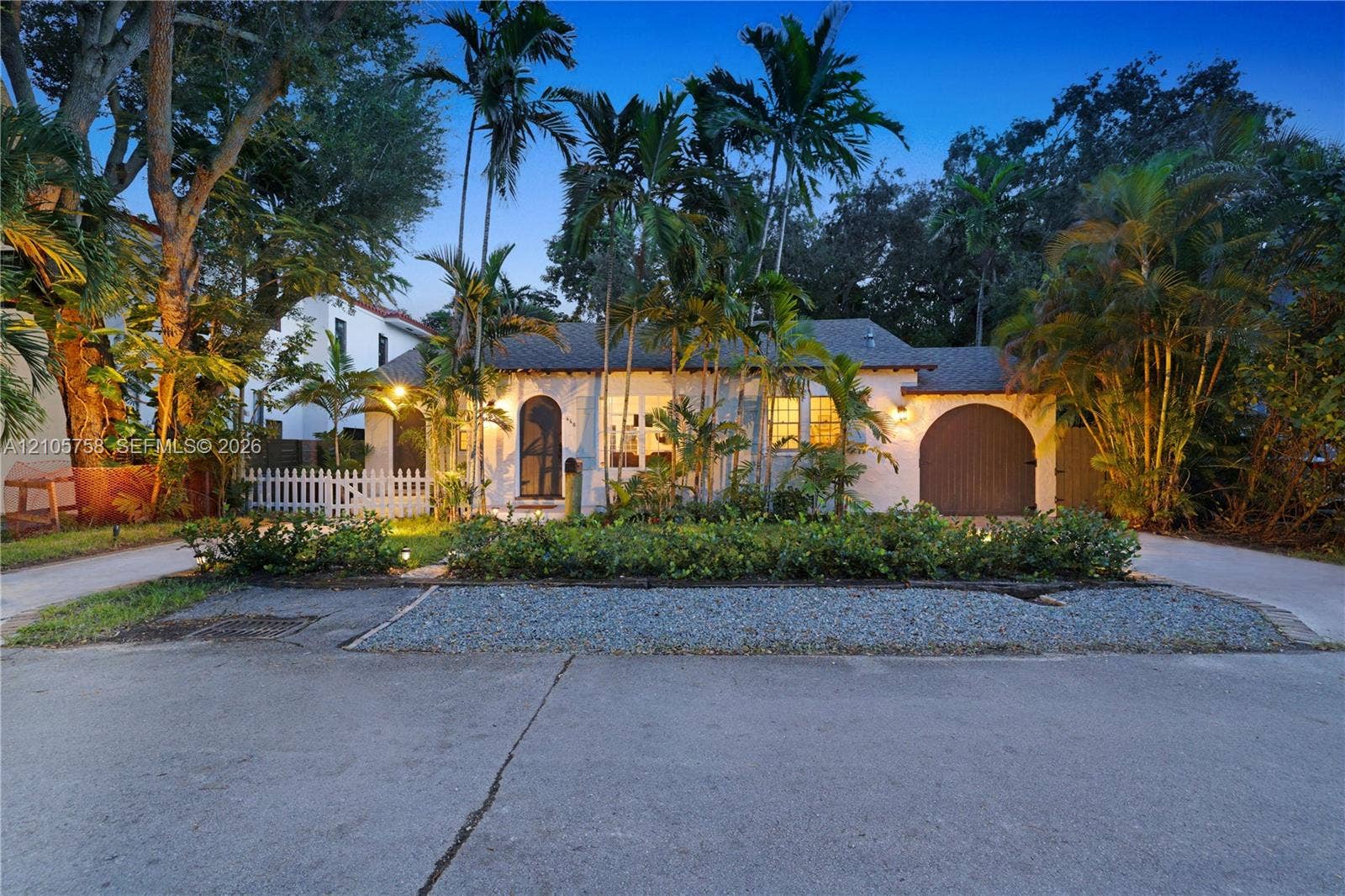 668 NE 70th Street, Miami, FL 33138 Stucco exterior residence featuring an arched entry, attached arched-top garage, and a picket fence, complemented by mature palm trees and established landscaping