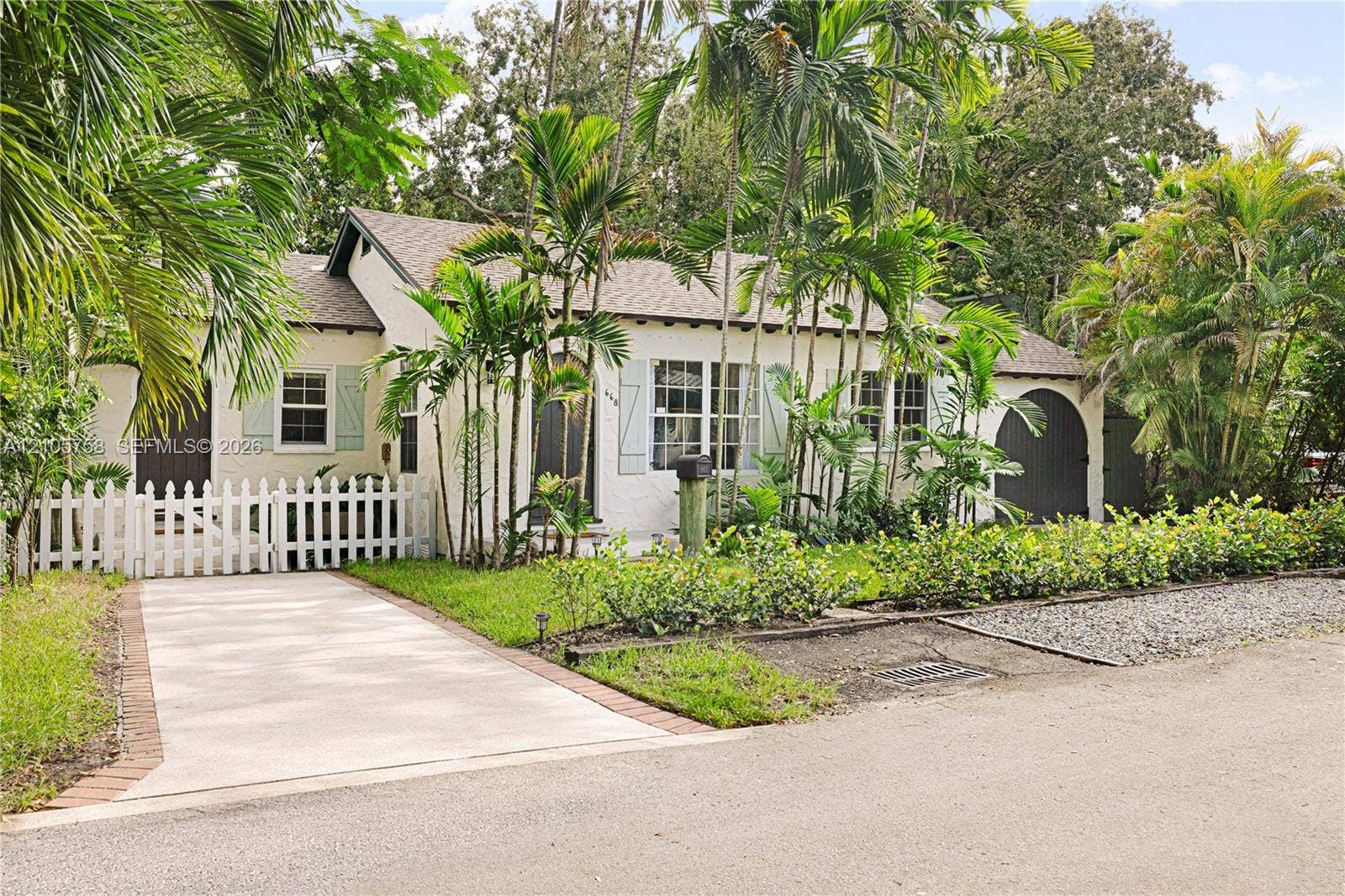 668 NE 70th Street, Miami, FL 33138 Stucco exterior featuring arched entry detailing, light-toned window shutters, a white picket fence, concrete driveway with brick edging, and mature palm landscaping