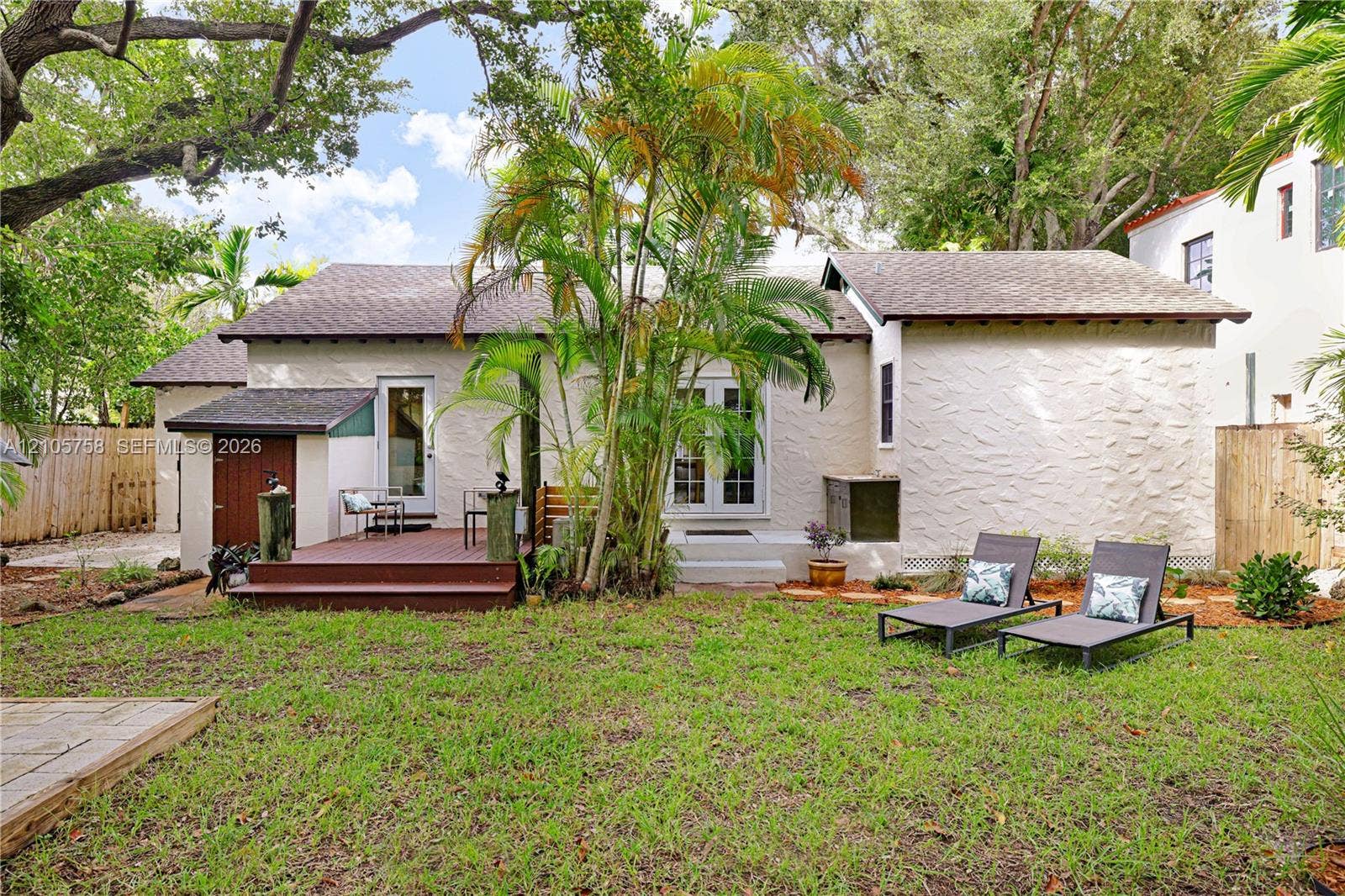 668 NE 70th Street, Miami, FL 33138 Stucco exterior featuring a raised wood-finish deck, glass-paneled double doors, mature palm trees, and a fenced backyard