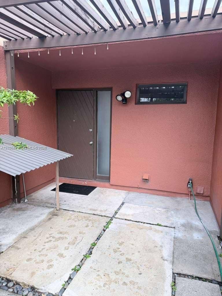 10343 Greenhouse Road, Pembroke Pines, FL 33026 Entryway featuring a structural pergola, diagonal-paneled door with a frosted glass sidelight, concrete patio, and exterior wall-mounted lighting