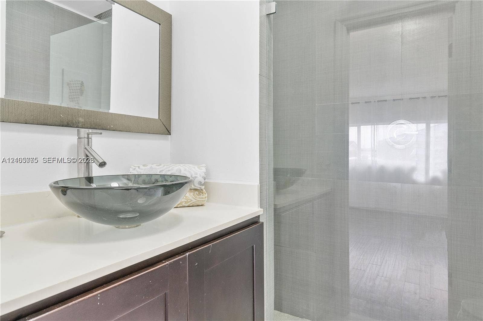 21230 NE 24th Court, Miami, FL 33180 Bathroom vanity featuring a dark-finish wood cabinet, solid-surface countertop, glass vessel sink, and a framed wall mirror, accompanied by a glass-enclosed shower with tiled surround