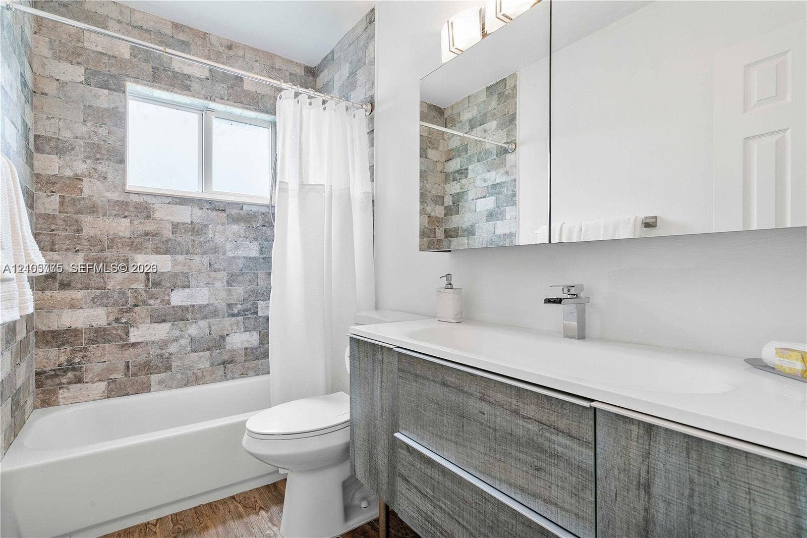 21230 NE 24th Court, Miami, FL 33180 Textured stone tile wall encompassing the bathtub, wood-finish floating vanity, integrated rectangular sink, wall-mounted faucet, and mirrored medicine cabinet
