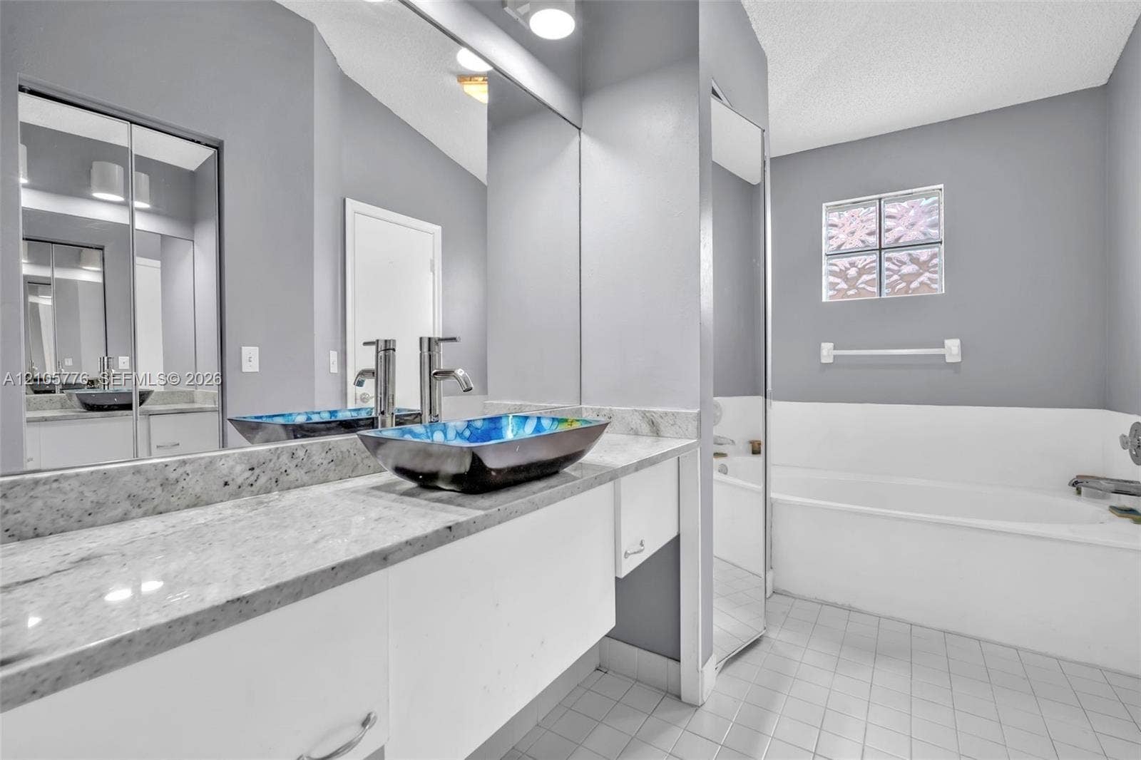 Stone-surface vanity with a decorative vessel sink, wall-spanning vanity mirror, floor-to-ceiling mirror panel, glass block window, and tile flooring
