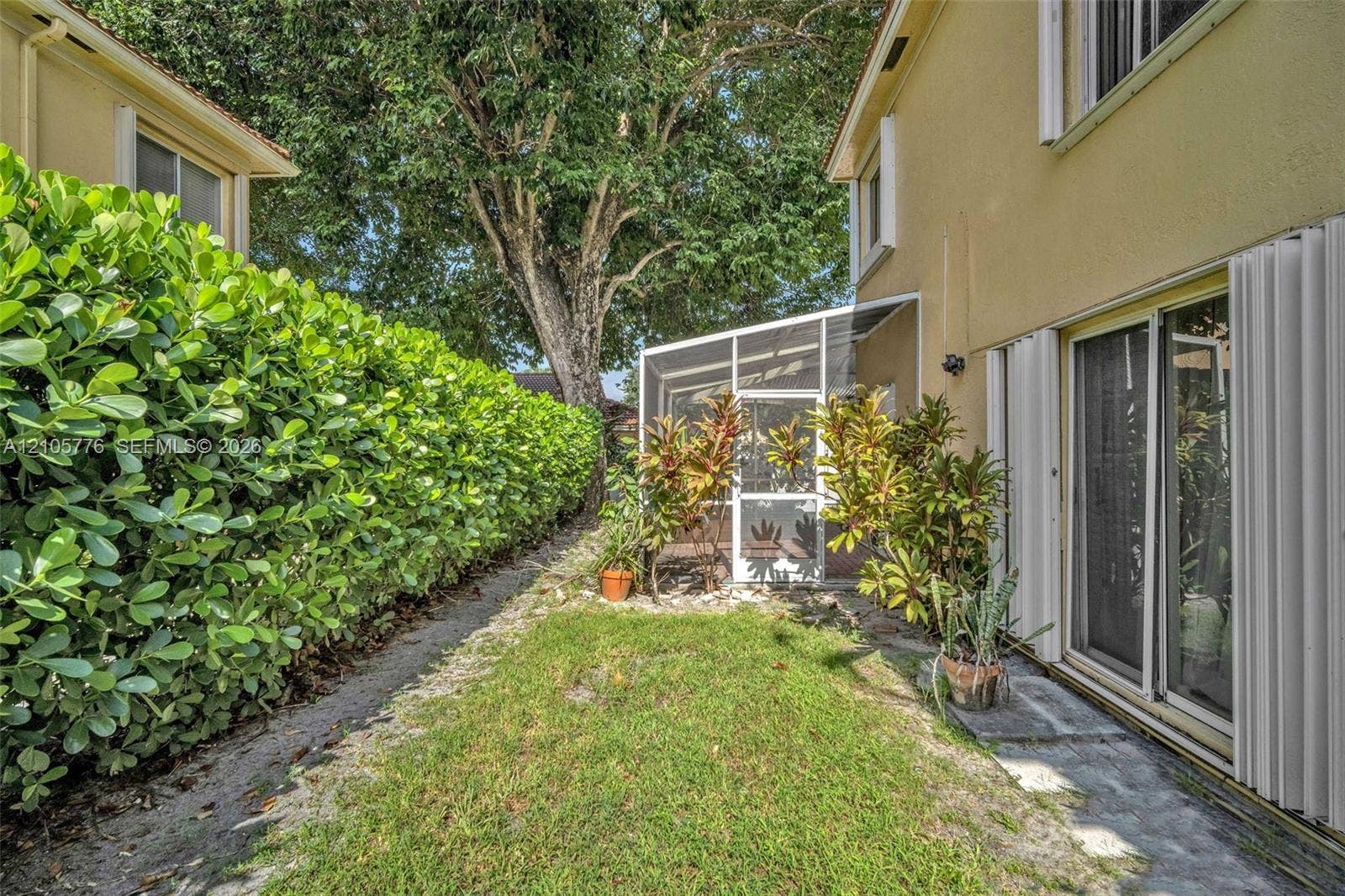 Exterior side yard featuring a manicured lawn, tall privacy hedge, screened-in enclosure, and sliding glass doors with accordion shutters