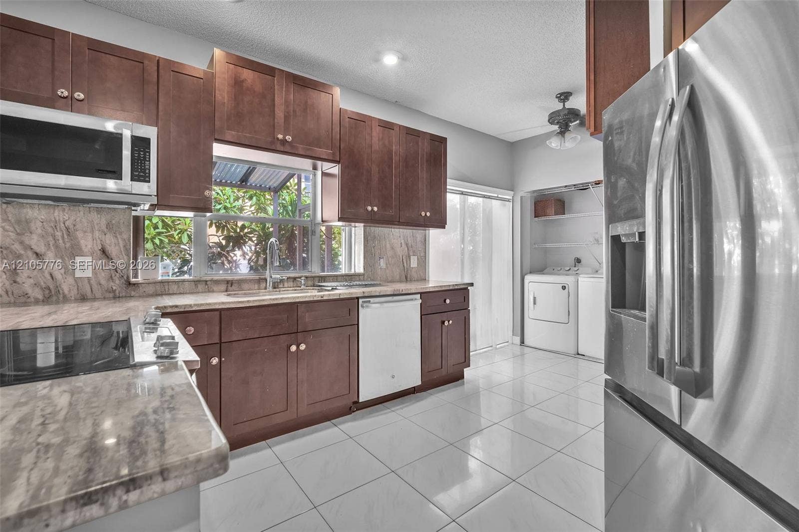 Kitchen featuring dark wood cabinetry, stone-patterned countertops, large-format tile flooring, stainless steel appliances, and a dedicated laundry area with built-in shelving