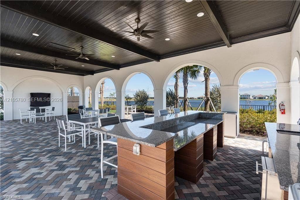 Covered outdoor kitchen featuring an expansive stone countertop, wood-paneled cabinetry, arched architectural pillars, a dark wood-finish plank ceiling, and herringbone patterned paver flooring