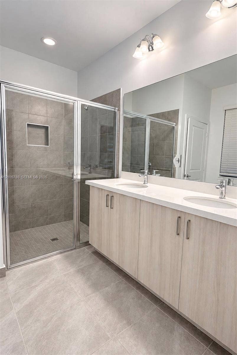 Dual-sink vanity with light wood-finish cabinetry, solid surface countertop, walk-in shower with full-height tile surround and built-in niche, and large vanity mirror