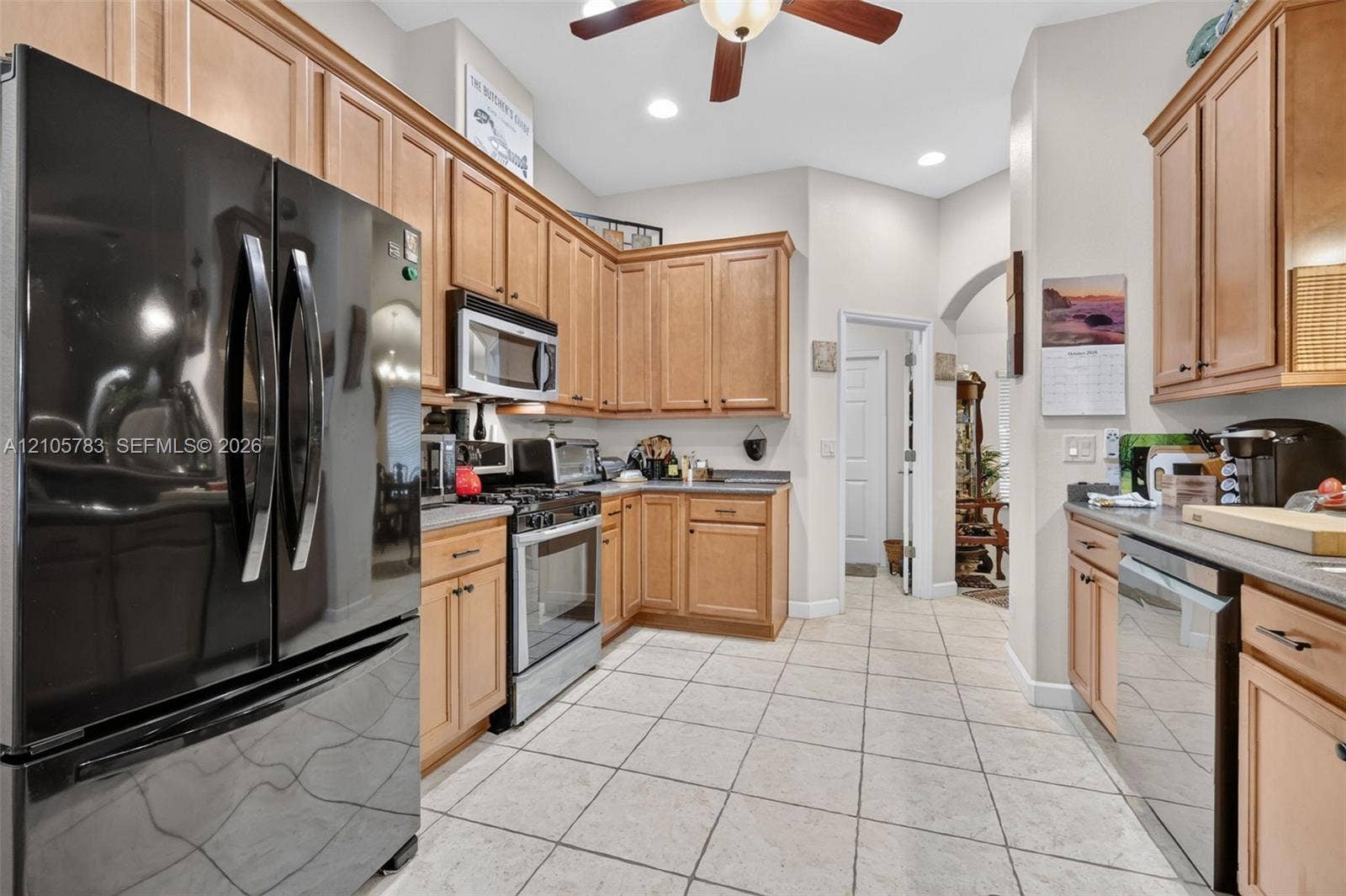 Kitchen featuring natural wood cabinetry, light-toned stone tile flooring, neutral countertops, stainless steel appliance suite, and a ceiling fan with integrated lighting