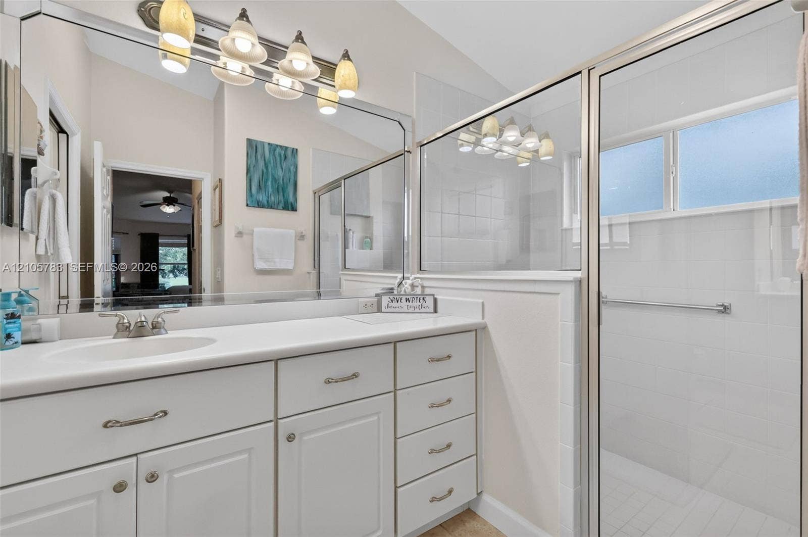 Bathroom vanity with white cabinetry, solid surface countertop, integrated sink, and large wall-mounted mirror, featuring a walk-in shower with tiled surround and glass enclosure