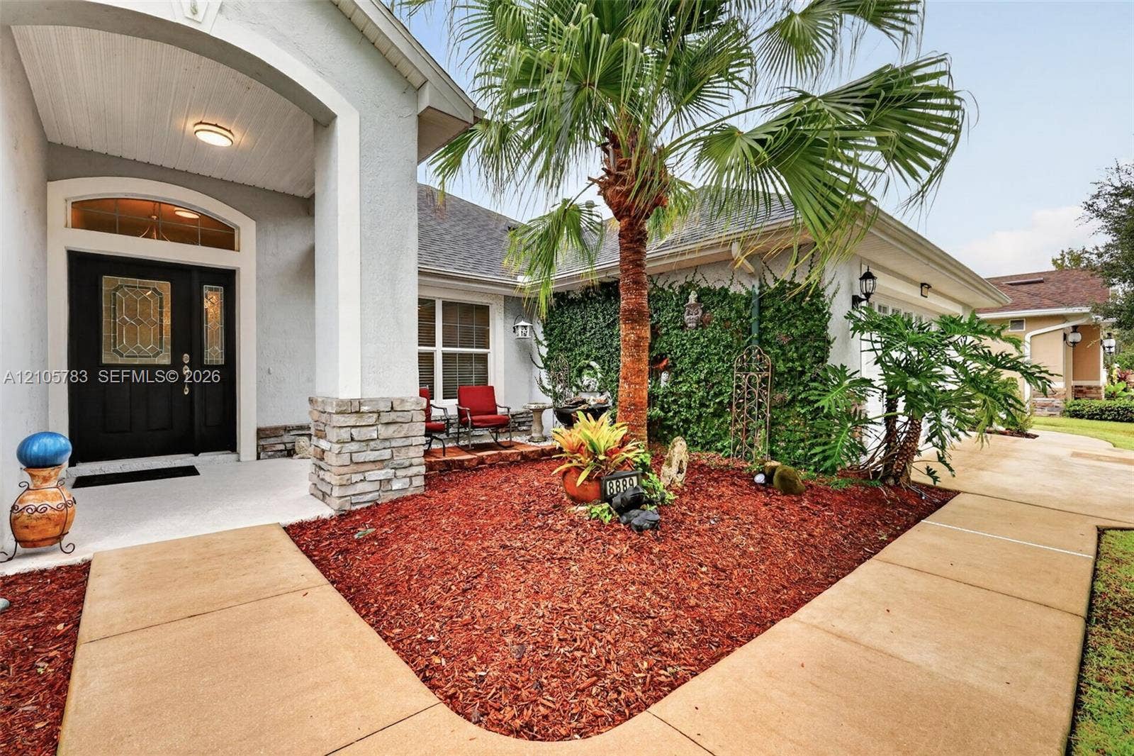 Covered entryway featuring a stone-accented column, recessed ceiling lighting, and a front door with decorative glass paneling, complemented by a front garden bed with mature palm landscaping