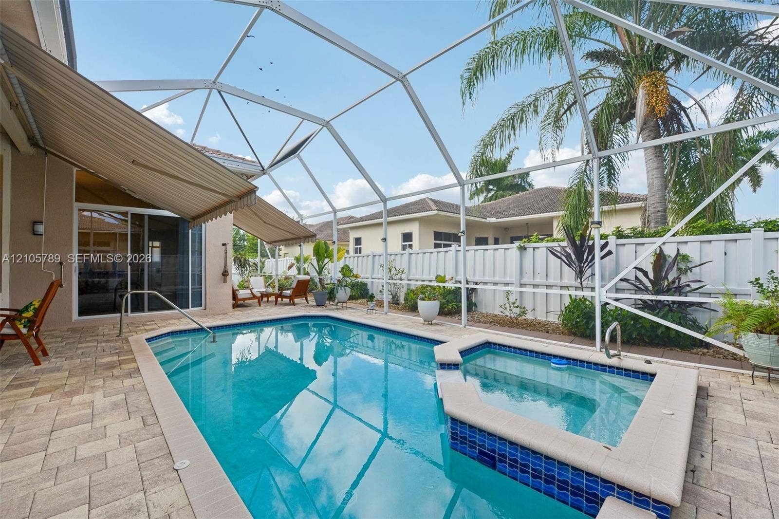 6353 SW 191st Avenue, Pembroke Pines, FL 33332 Screen-enclosed pool area featuring a spillover spa with blue tile accents, paver patio, retractable awning, and perimeter privacy fencing