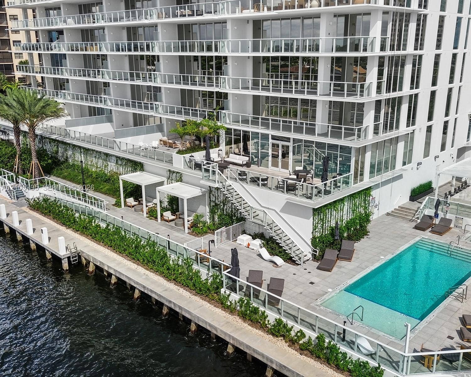 407 N Riverside Drive, #202, Pompano Beach, Pompano Beach, FL 33062 High-rise building exterior featuring tiered balconies with glass railings, a waterfront pool deck, stone-paved sun terrace, integrated pergolas, and vertical landscaping