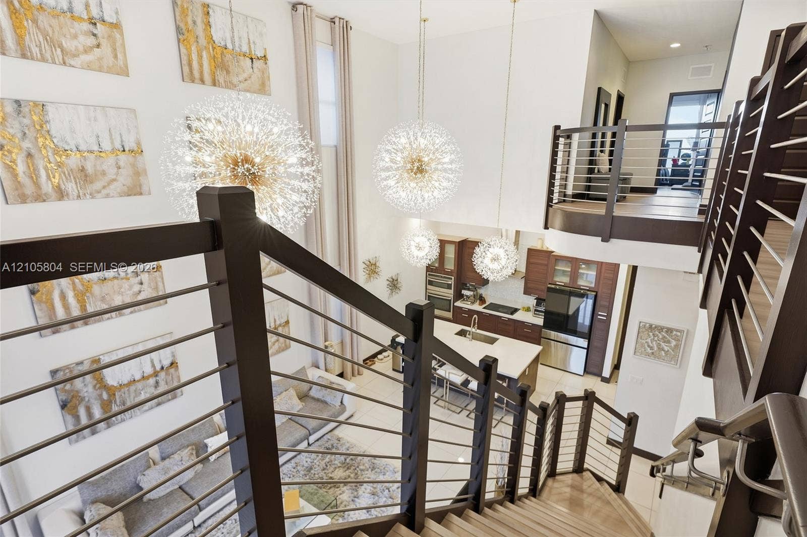 15932 SW 136th Way, Miami, FL 33196 Double-height great room featuring multi-tiered sputnik-style chandeliers, metal cable stair railings, and an open-concept kitchen with built-in cabinetry and stone-surfaced island