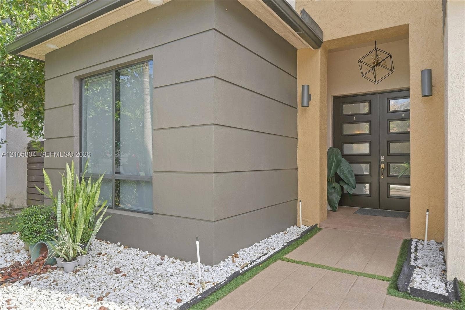 15932 SW 136th Way, Miami, FL 33196 Modern exterior facade featuring smooth stucco finishes, horizontal architectural detailing, double-entry door with glass panels, geometric metal pendant lighting, and a paved stone walkway
