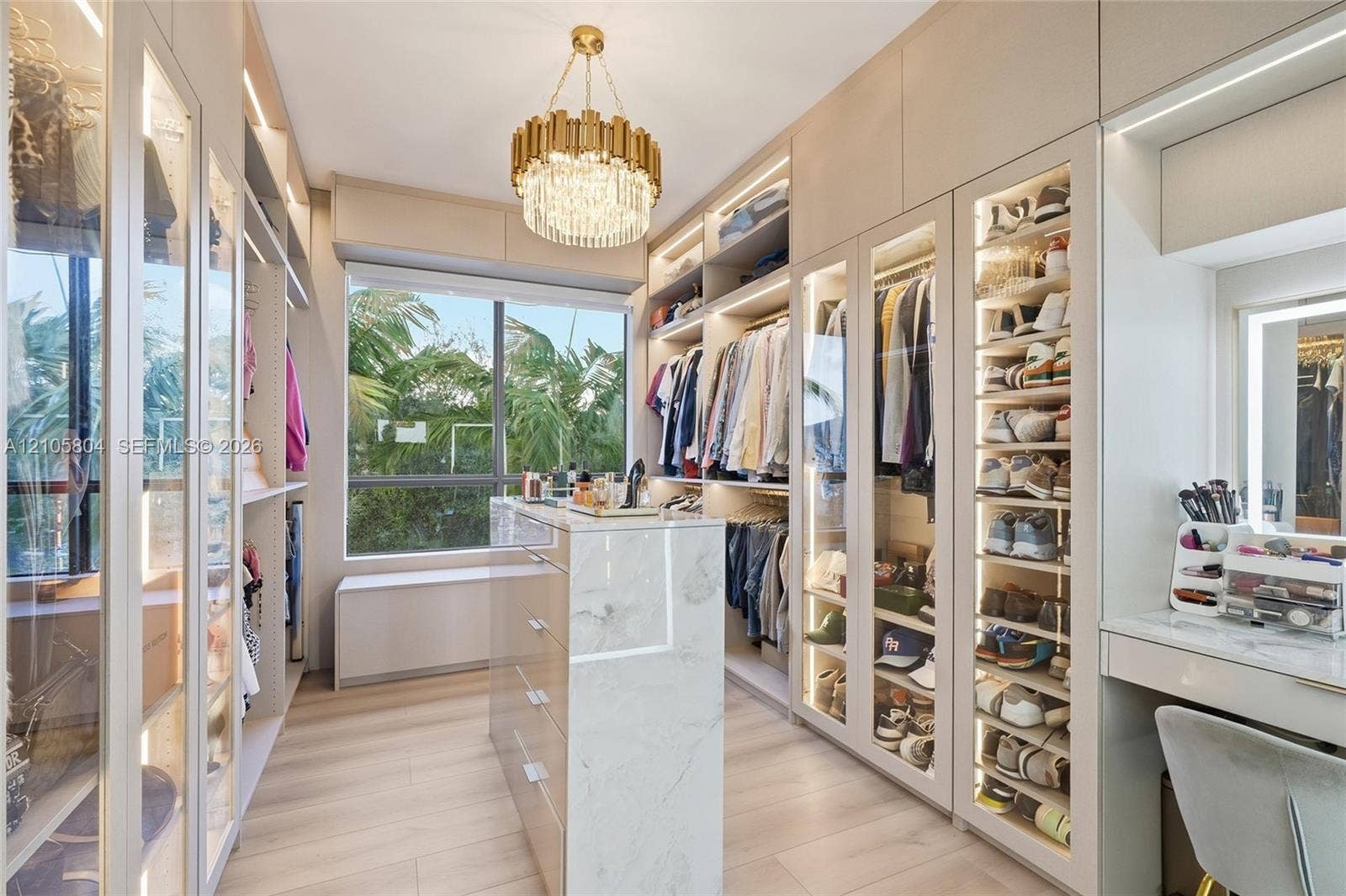 15932 SW 136th Way, Miami, FL 33196 Custom walk-in closet featuring floor-to-ceiling glass-front cabinetry, integrated LED lighting, stone-finish center island, light wood-finish flooring, and a dedicated vanity station