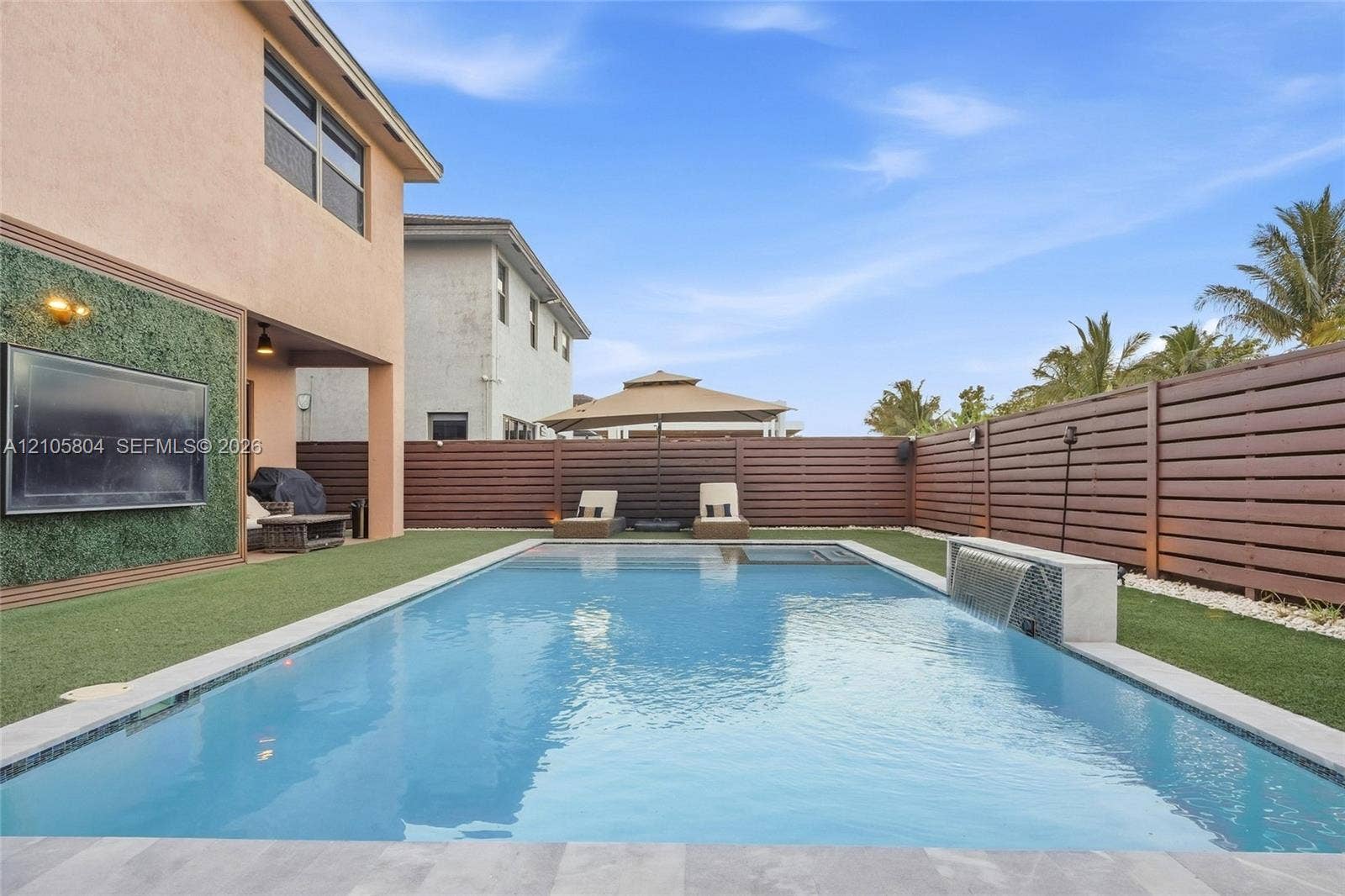 15932 SW 136th Way, Miami, FL 33196 Backyard featuring a rectangular swimming pool with a built-in water feature, horizontal wood-slat privacy fencing, artificial turf lawn, and a wall-mounted exterior television