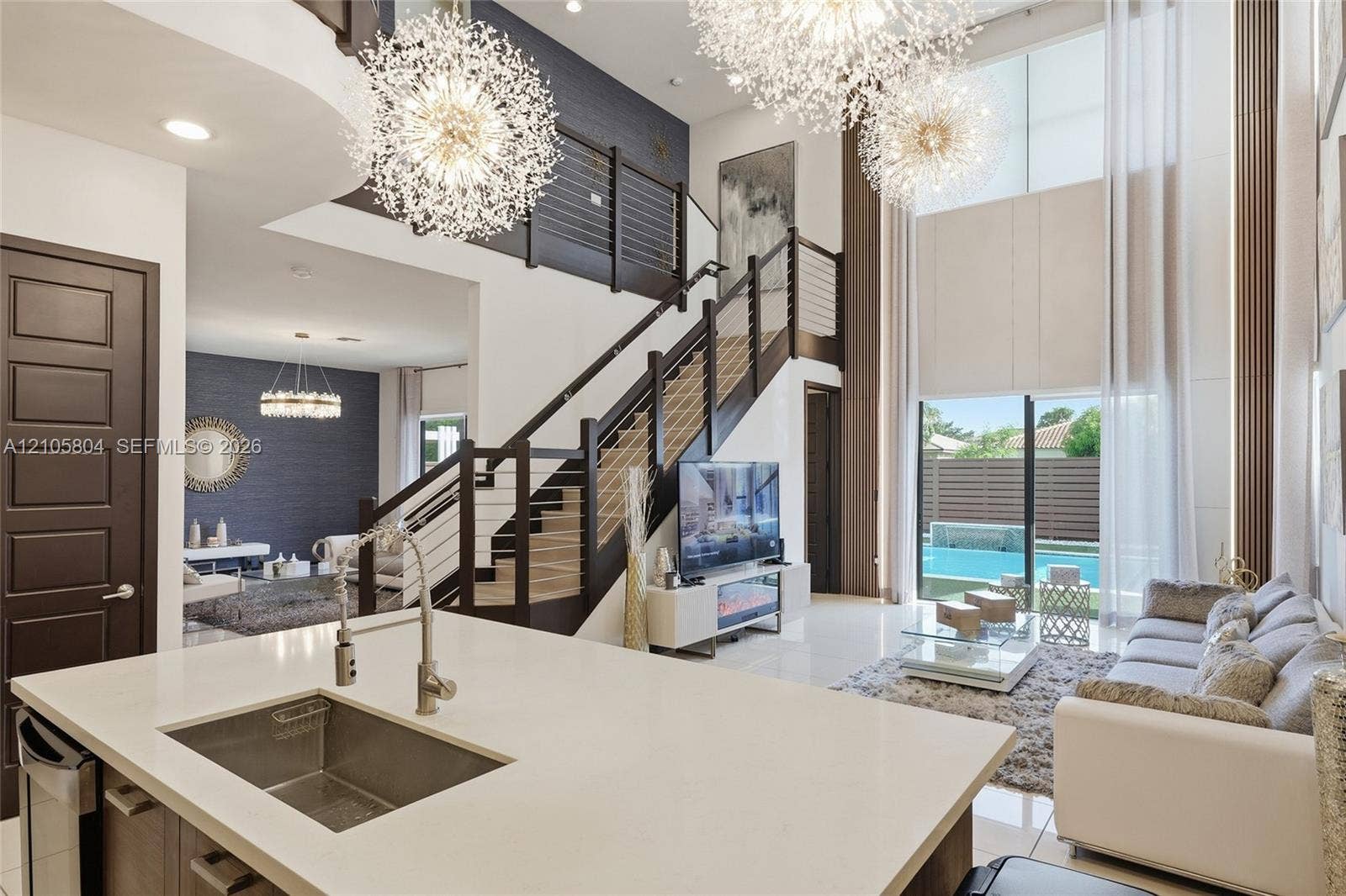 15932 SW 136th Way, Miami, FL 33196 Double-height ceiling, open-plan interior featuring a floating staircase with cable railing, expansive floor-to-ceiling windows, dual sputnik-style chandeliers, and a kitchen island with a stone-finish countertop