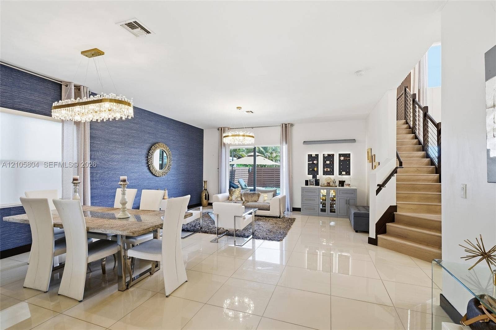 15932 SW 136th Way, Miami, FL 33196 Open-concept interior featuring oversized tile flooring, a blue textured accent wall, dual crystal chandeliers, and a staircase with wood-tread stairs and a metal cable railing system
