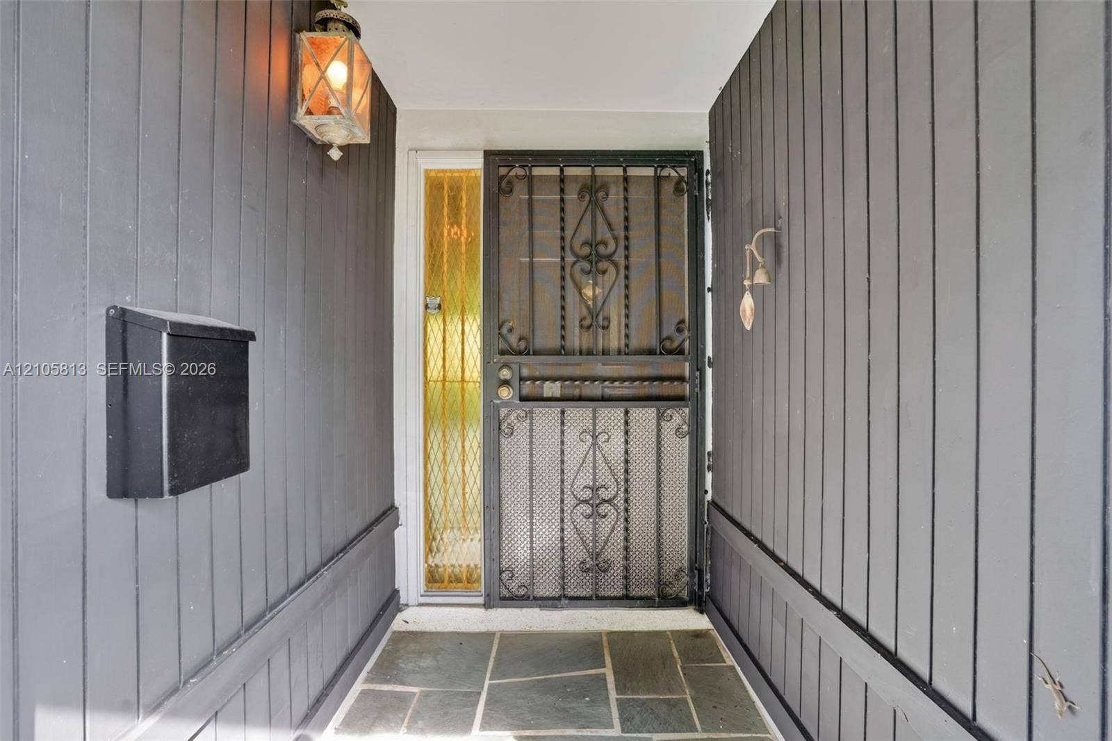 Covered entryway featuring vertical dark-tone wood siding, flagstone flooring, a decorative metal security door, a sidelight with textured glass, and a wall-mounted lantern-style fixture