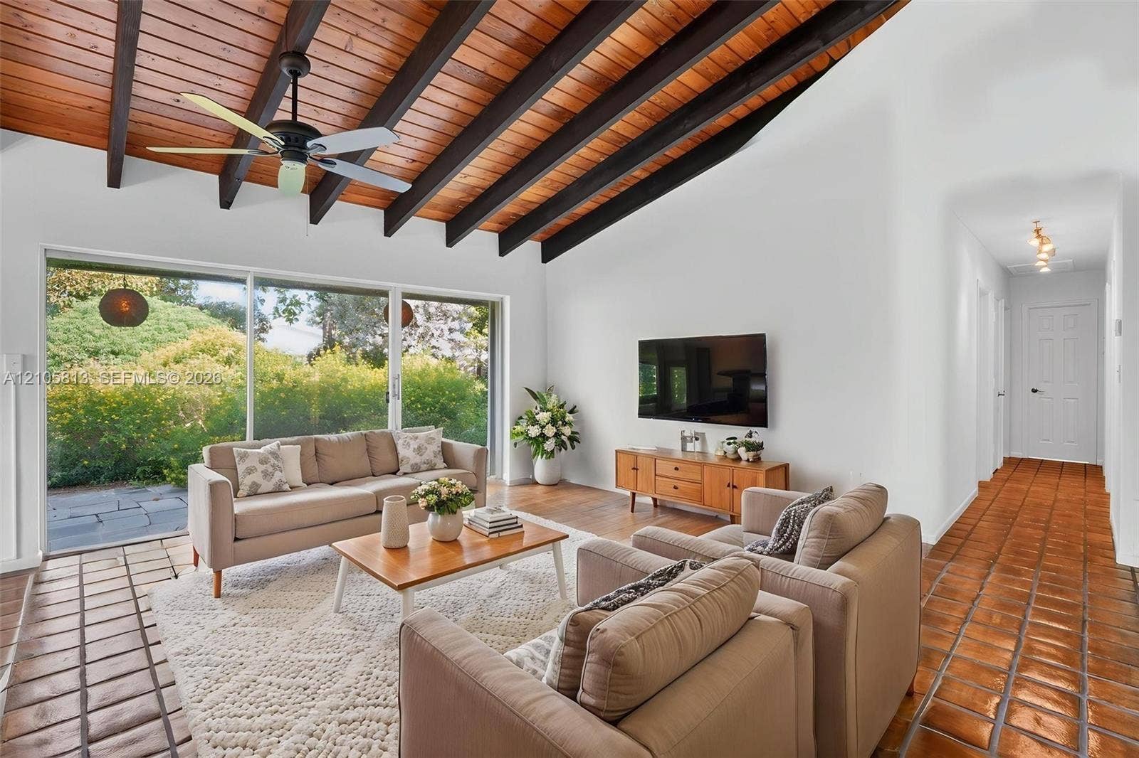 Vaulted ceiling featuring exposed wood beams, stained wood planking, and a central ceiling fan, complemented by extensive floor-to-ceiling glass sliding doors and terracotta tile flooring