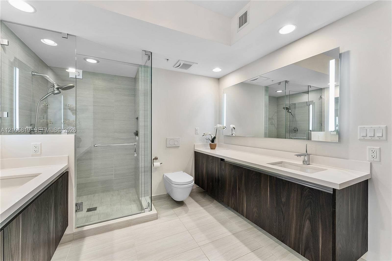 1180 N Federal Highway, #810, Fort Lauderdale, FL 33304 Floating dark wood-finish dual vanity with solid surface countertops, frameless glass-enclosed shower, wall-mounted toilet, and large integrated vanity mirror with LED lighting