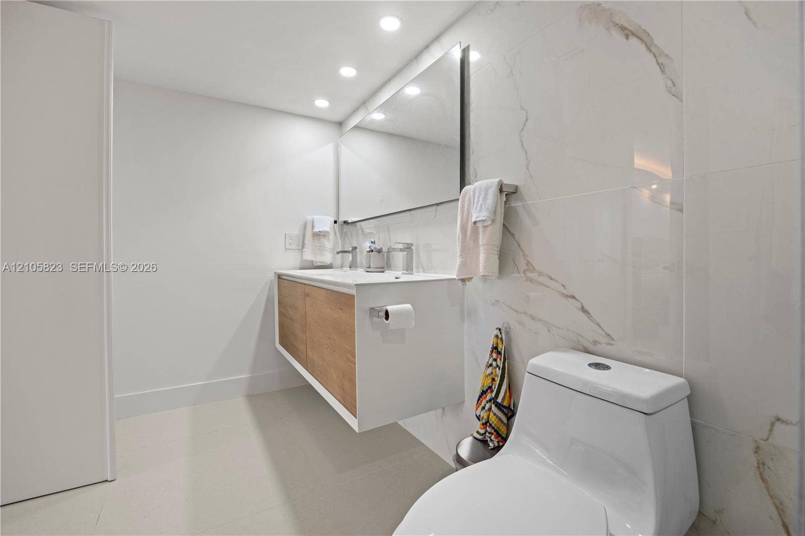 Floating vanity with wood-finish cabinetry, rectangular vessel sink, large wall-mounted mirror, recessed ceiling lighting, and floor-to-ceiling porcelain wall tile