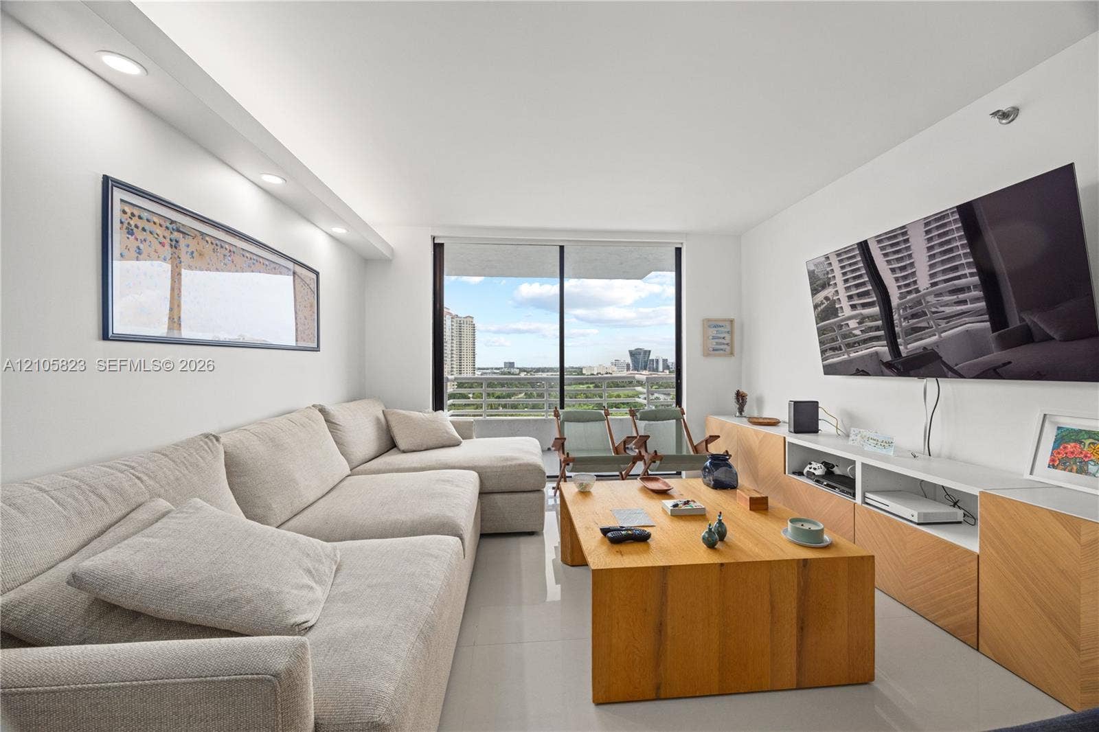 Living area featuring light-toned tile flooring, recessed ceiling lighting, built-in wooden media cabinetry with a chevron pattern, and floor-to-ceiling glass sliding doors opening to an exterior balcony