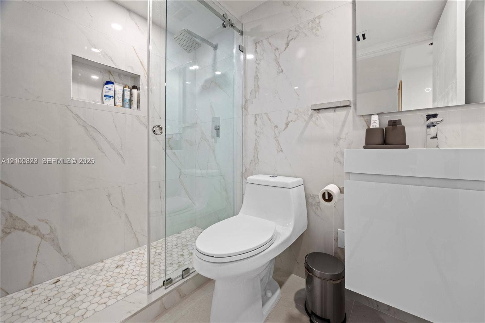 Bathroom featuring wall-to-wall large-format porcelain tile, glass-enclosed shower with integrated shelving, hexagonal floor tiling, and wall-mounted vanity with a minimalist faucet