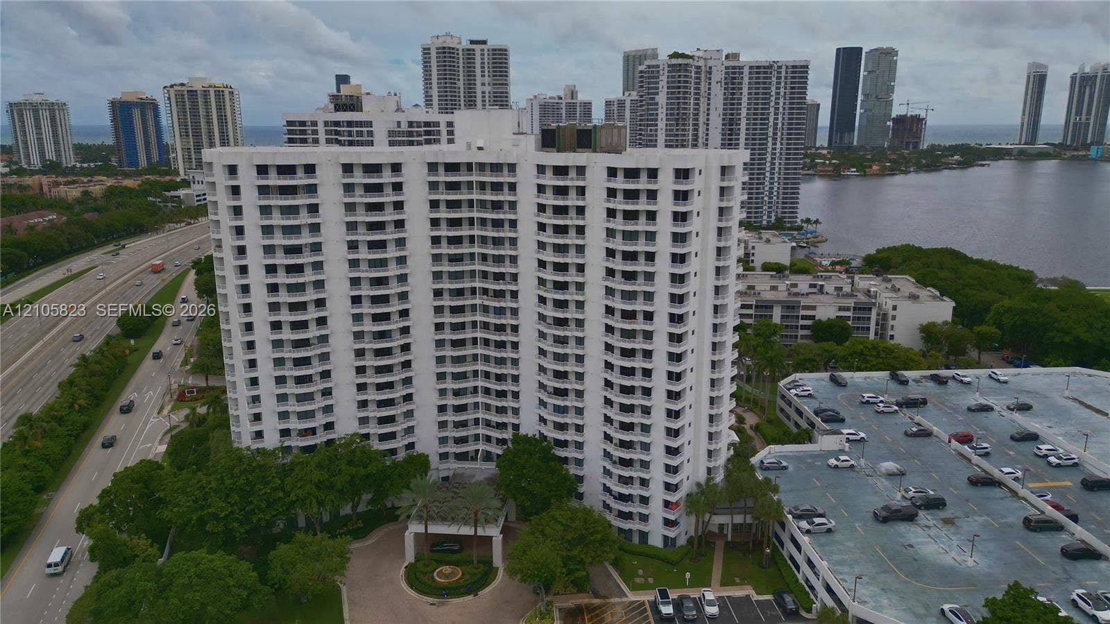 High-rise residential building featuring stacked private balconies, a covered porte-cochre entrance, paved circular driveway, and an attached multi-level parking structure