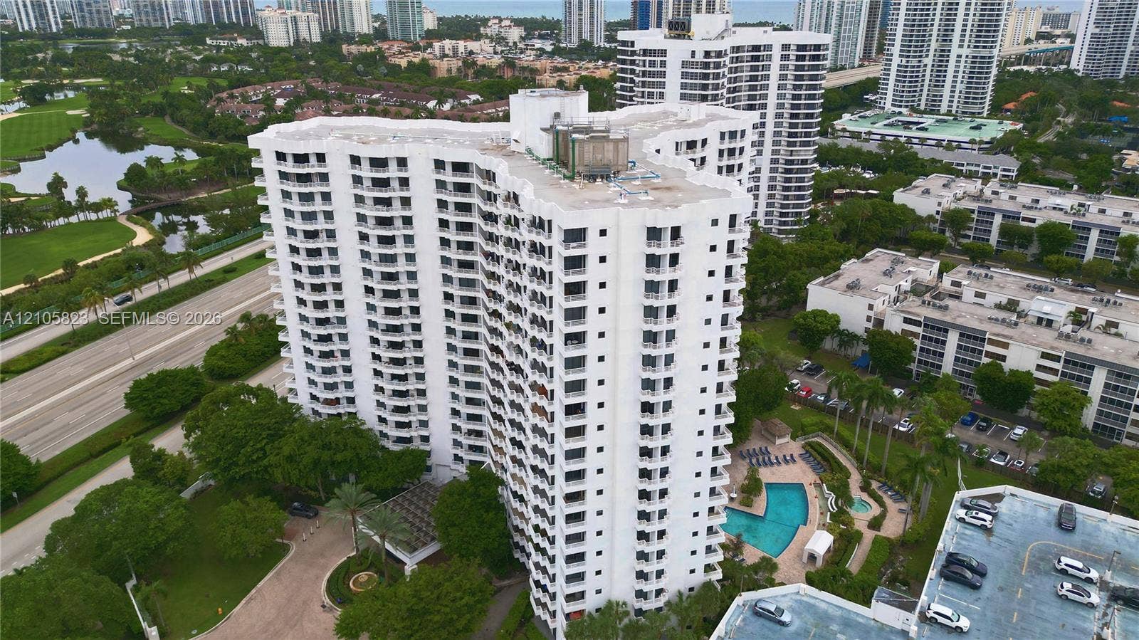 High-rise residential building featuring expansive private balconies, a ground-level swimming pool, manicured landscaping, and structured parking