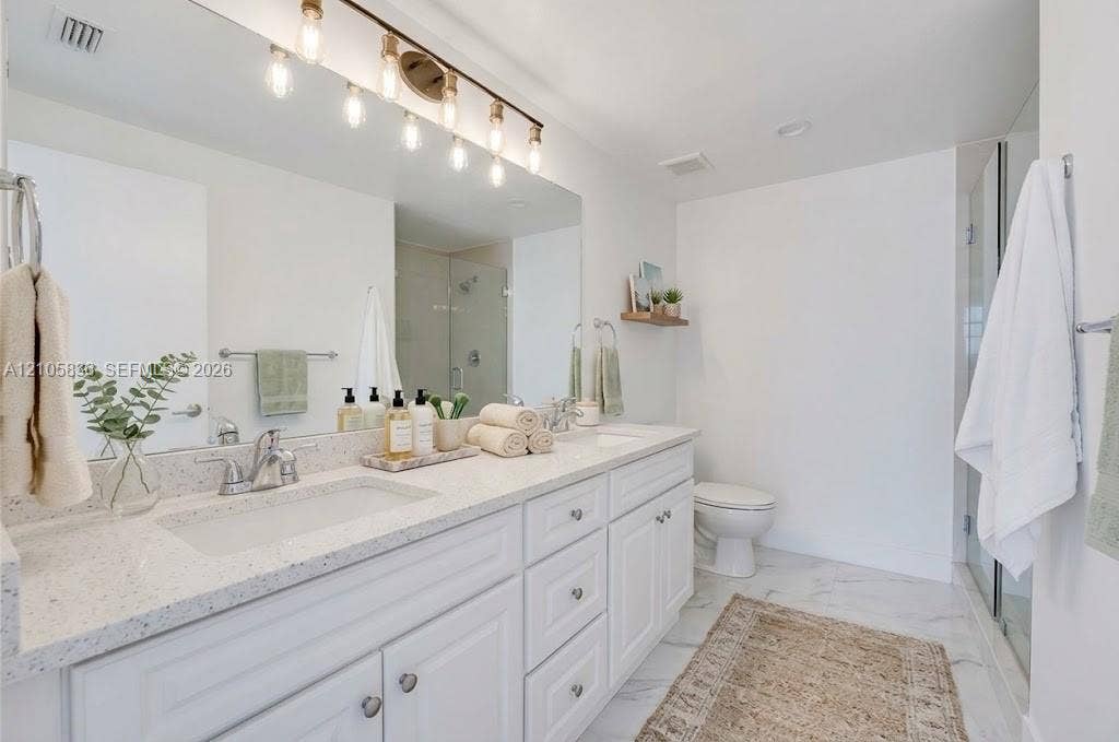 2475 Brickell Avenue, #704, Miami, FL 33129 Dual-sink vanity with white cabinetry, light-toned stone-finish countertops, polished chrome fixtures, and a large-scale mirror with an integrated multi-bulb light fixture