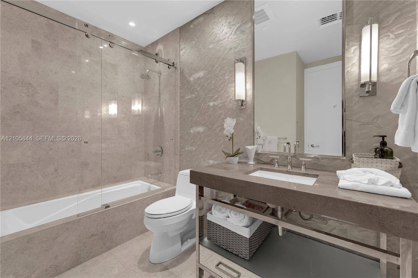 Bal Harbour, Bal Harbour, FL 33154 Stone-tiled bathtub enclosure with a frameless glass sliding door, stone-topped vanity with an integrated basin, metal-framed open base, and vertically mounted wall sconces
