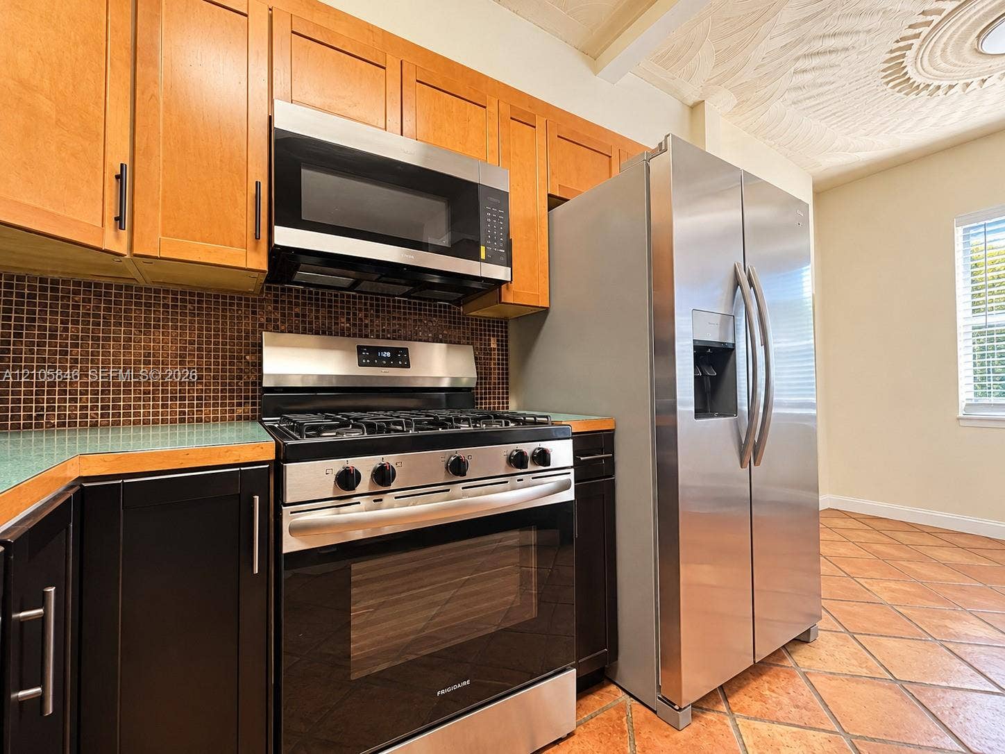 Kitchen featuring two-tone cabinetry, stainless steel gas range and refrigerator, mosaic tile backsplash, and terracotta tile flooring