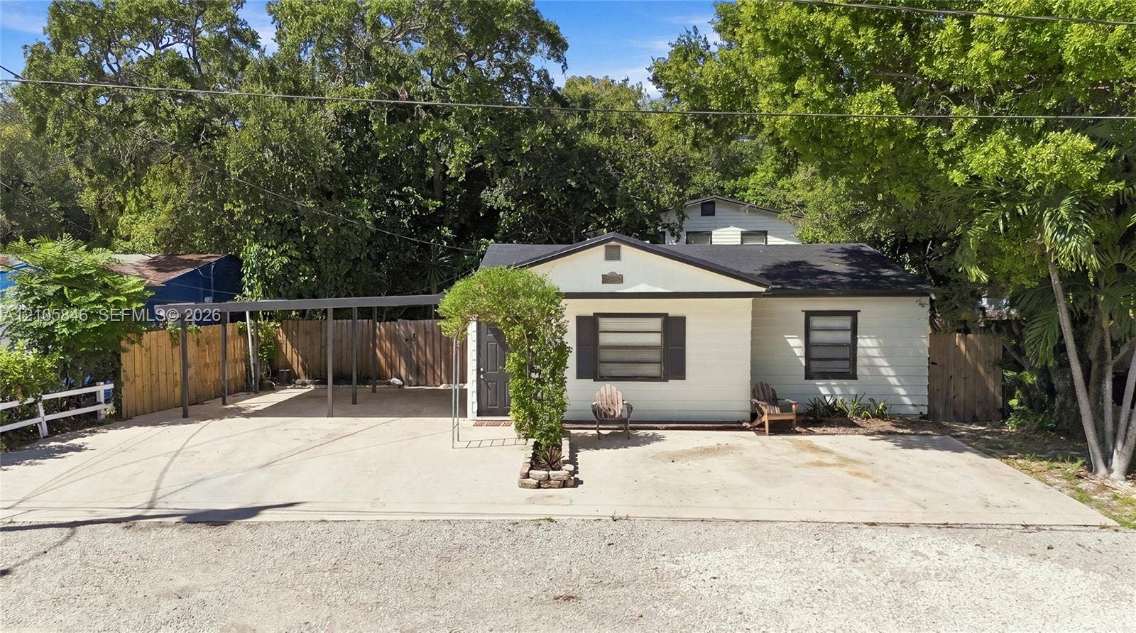 Single-story residence featuring light-colored horizontal siding, dark-toned shutters, an attached metal-roofed carport, a spacious concrete driveway, and surrounding mature trees