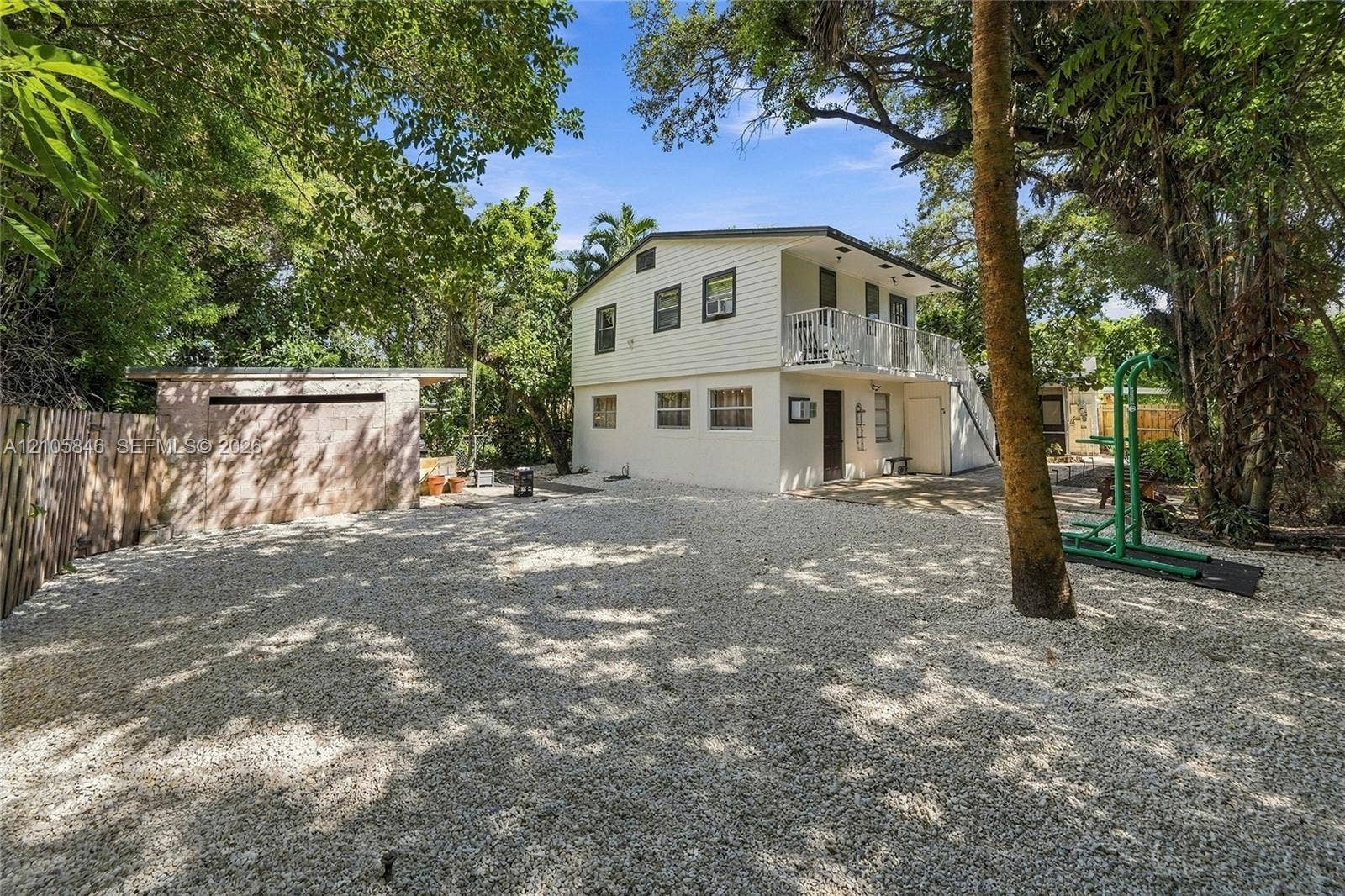 Two-story exterior with horizontal siding, gravel ground cover, detached concrete block structure, second-level balcony with metal railing, and mature trees