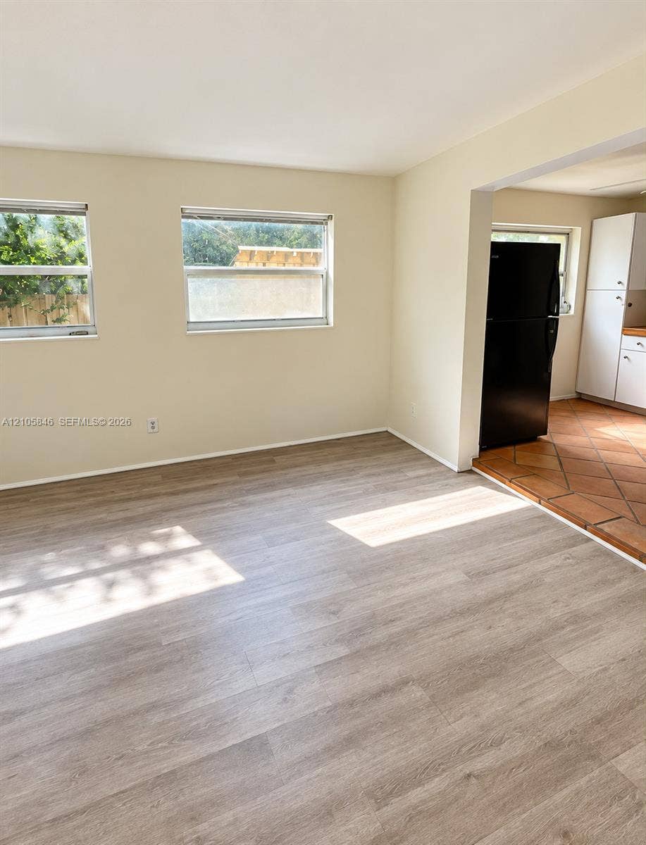 Neutral interior space featuring wood-finish flooring, dual windows, and terra-cotta tile flooring in the kitchen area with white cabinetry and a black refrigerator