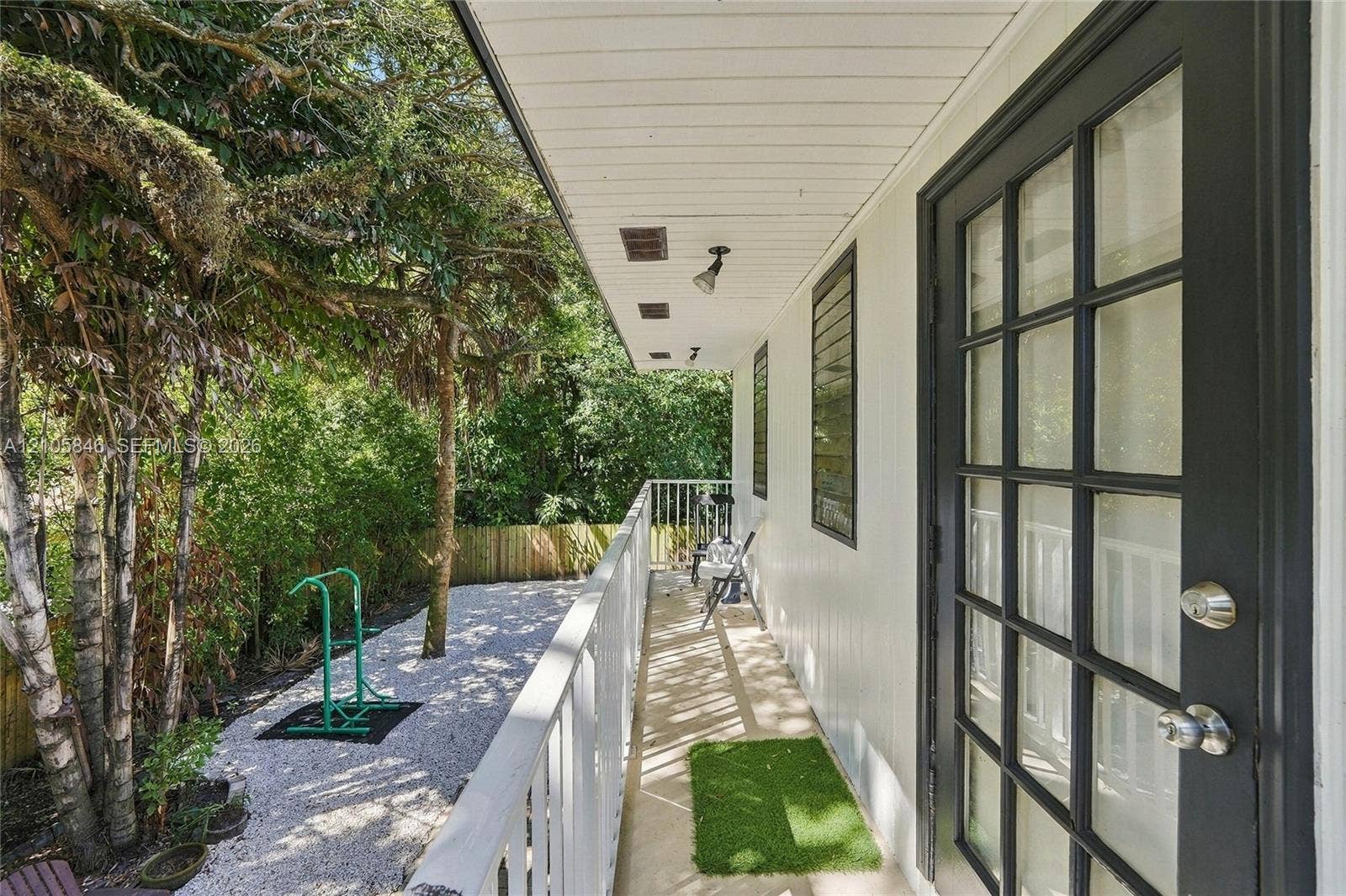 Covered exterior balcony featuring a white metal railing, black-framed French door, exterior wall-mounted lighting, and gravel ground cover