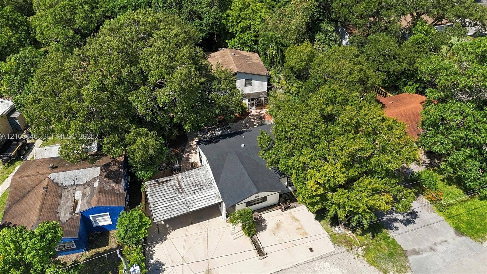 Aerial view featuring a multi-level structure with a dark shingled roof, a concrete driveway, and an attached covered carport, situated among mature trees