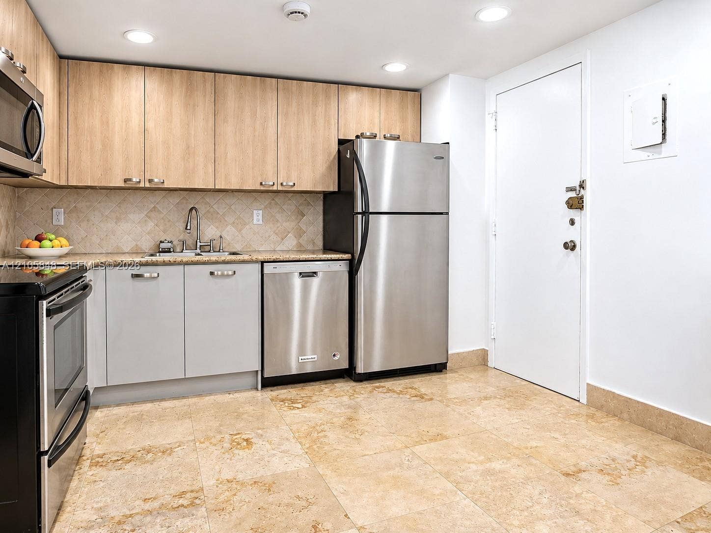 Kitchen featuring light wood-finish upper cabinetry, lower gray cabinetry, stone-tile flooring, stainless steel appliances, and a tiled backsplash