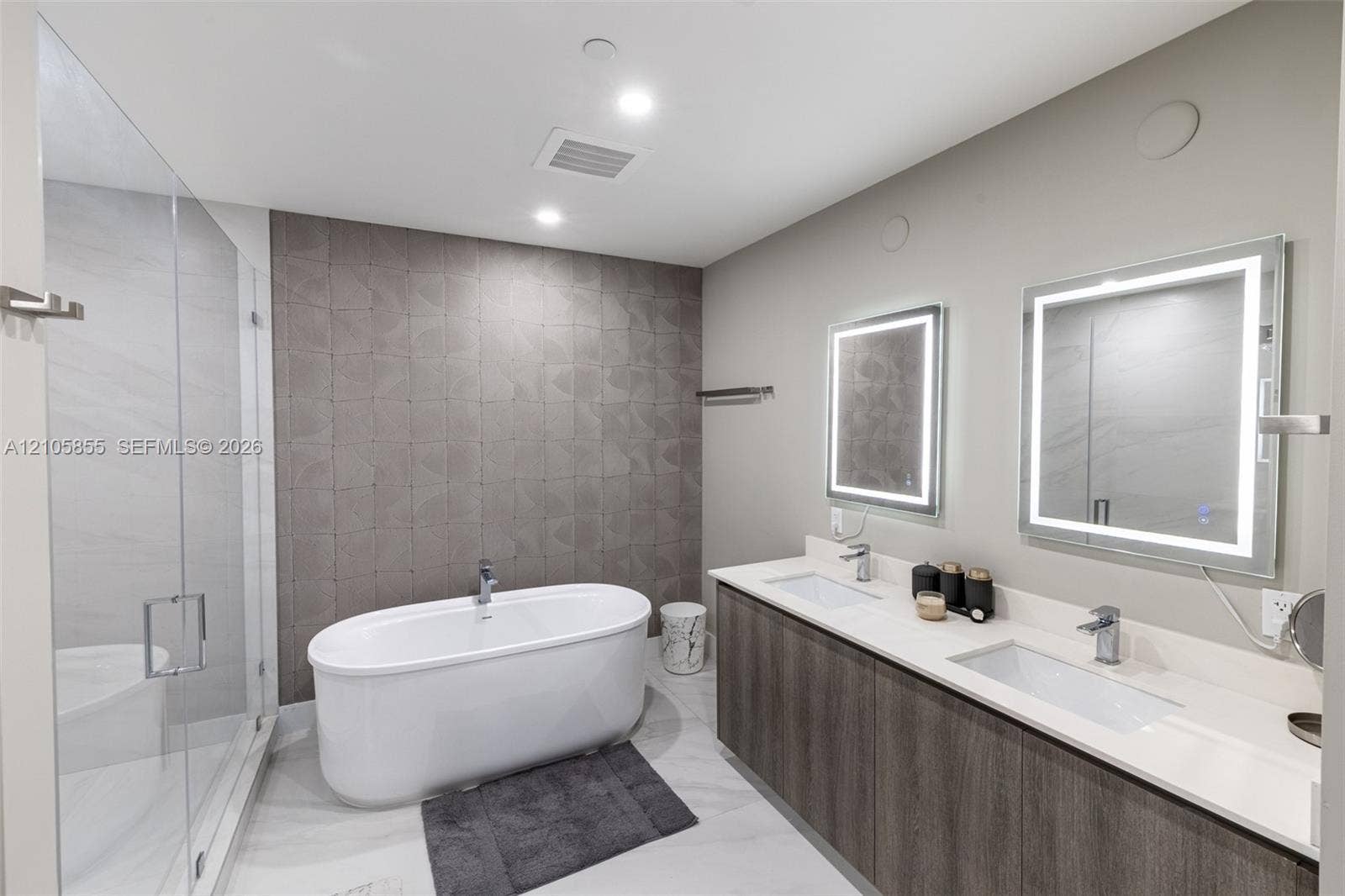 17550 Collins Avenue, #703, Sunny Isles Beach, Sunny Isles Beach, FL 33160 Freestanding soaking tub with a floor-mounted faucet, walk-in glass shower, dual-sink vanity with light-toned stone countertop, wood-finish cabinetry, and LED-backlit mirrors