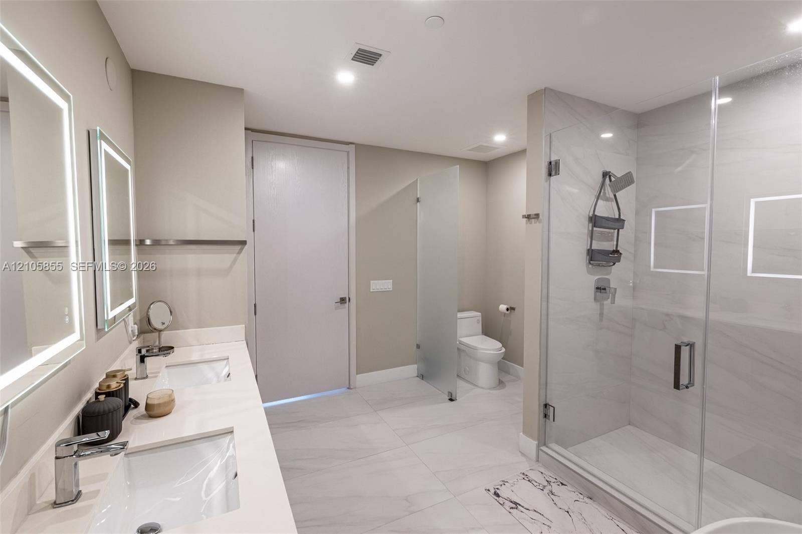 17550 Collins Avenue, #703, Sunny Isles Beach, Sunny Isles Beach, FL 33160 Dual-sink vanity with a solid-surface countertop and integrated lighting, walk-in shower with stone-style wall tile and recessed shelving, and a glass-enclosed water closet