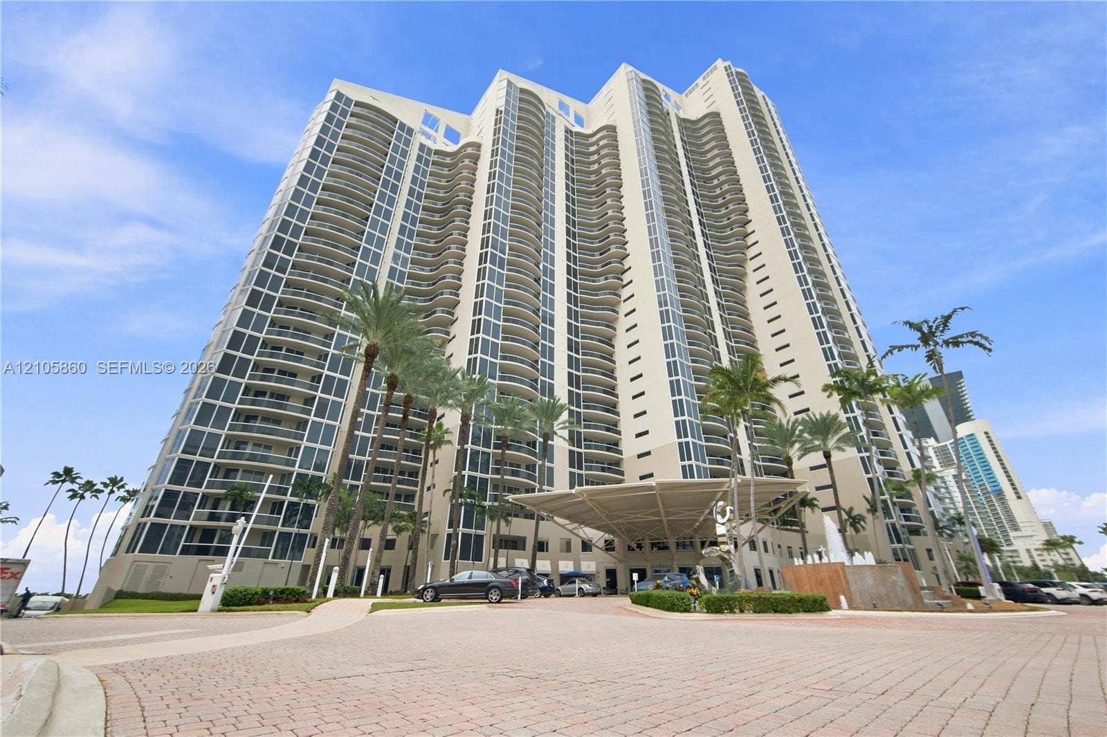 17555 Collins Avenue, #1903, Sunny Isles Beach, FL 33160 High-rise residential tower featuring a concrete facade, floor-to-ceiling windows, private balconies, a porte-cochre entry, and landscaped grounds with palm trees
