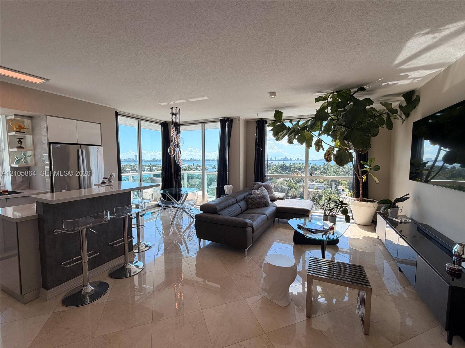 Open-concept living space featuring floor-to-ceiling windows, polished stone flooring, stainless steel kitchen appliances, and a breakfast bar with stone-finish surfacing