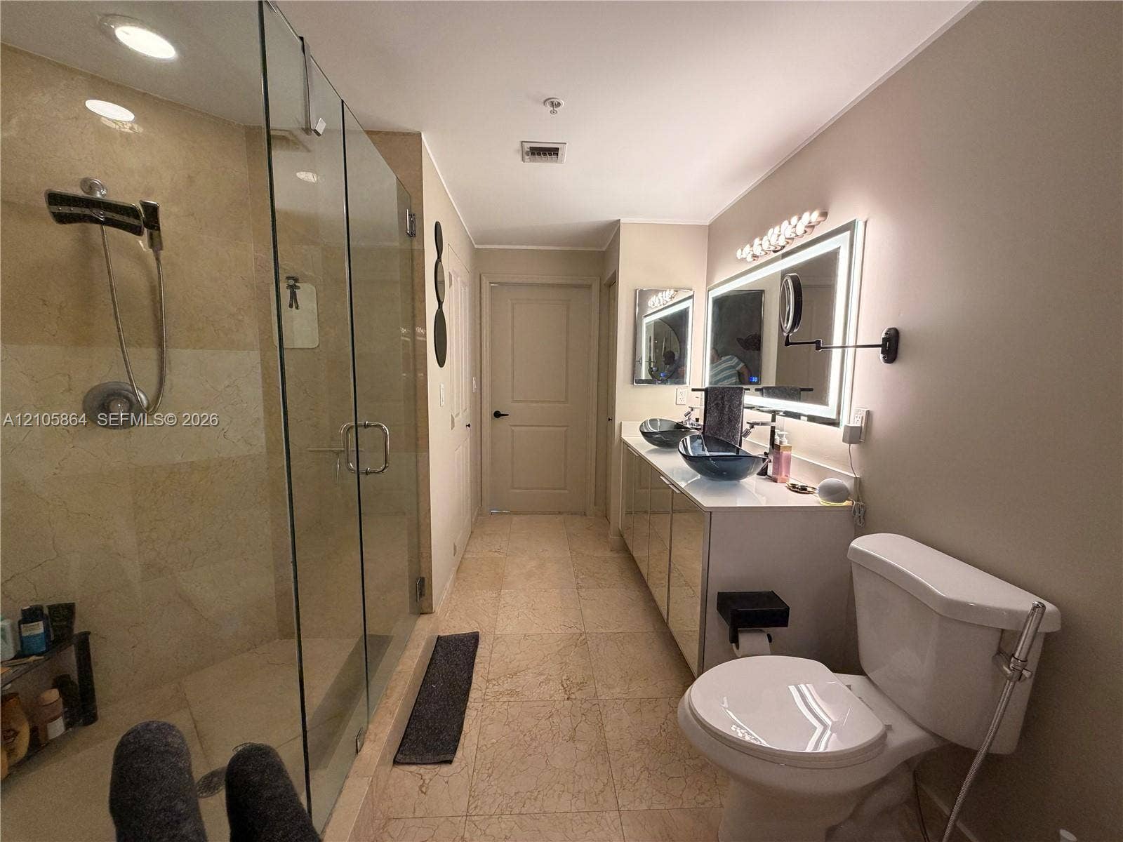 Bathroom featuring stone-tile flooring, glass-enclosed shower with stone wall surfacing, dual vanity with vessel sinks, backlit wall-mounted mirror, and wall-mounted articulating vanity mirror