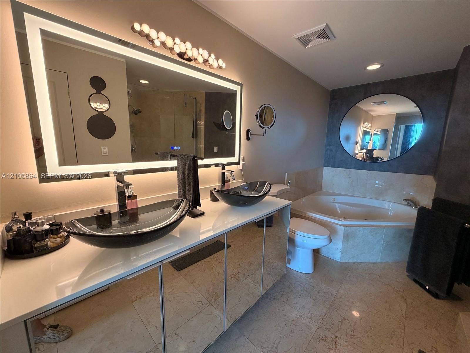 Double vanity featuring integrated backlighting, rectangular vessel sinks, stone countertop, corner jetted tub, and stone tile flooring