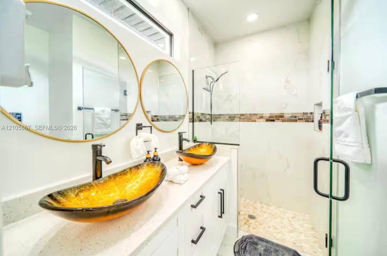 Dual vanity featuring white cabinetry, light-toned stone-finish countertops, vessel sinks, round mirrors with metallic frames, and a walk-in shower with stone tile flooring