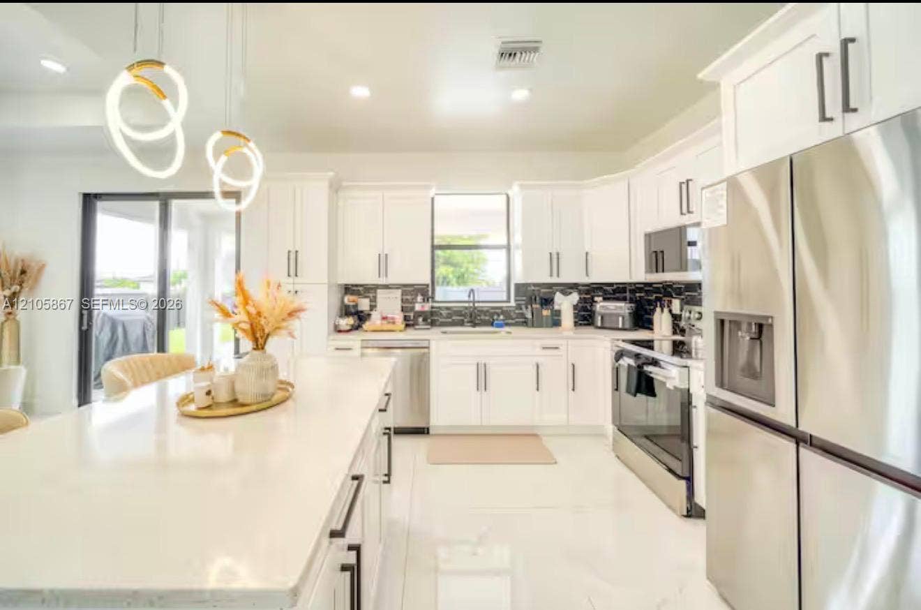 Kitchen featuring white shaker-style cabinetry, a quartz waterfall island, stainless steel appliances, a dark tile backsplash, and contemporary sculptural pendant lighting