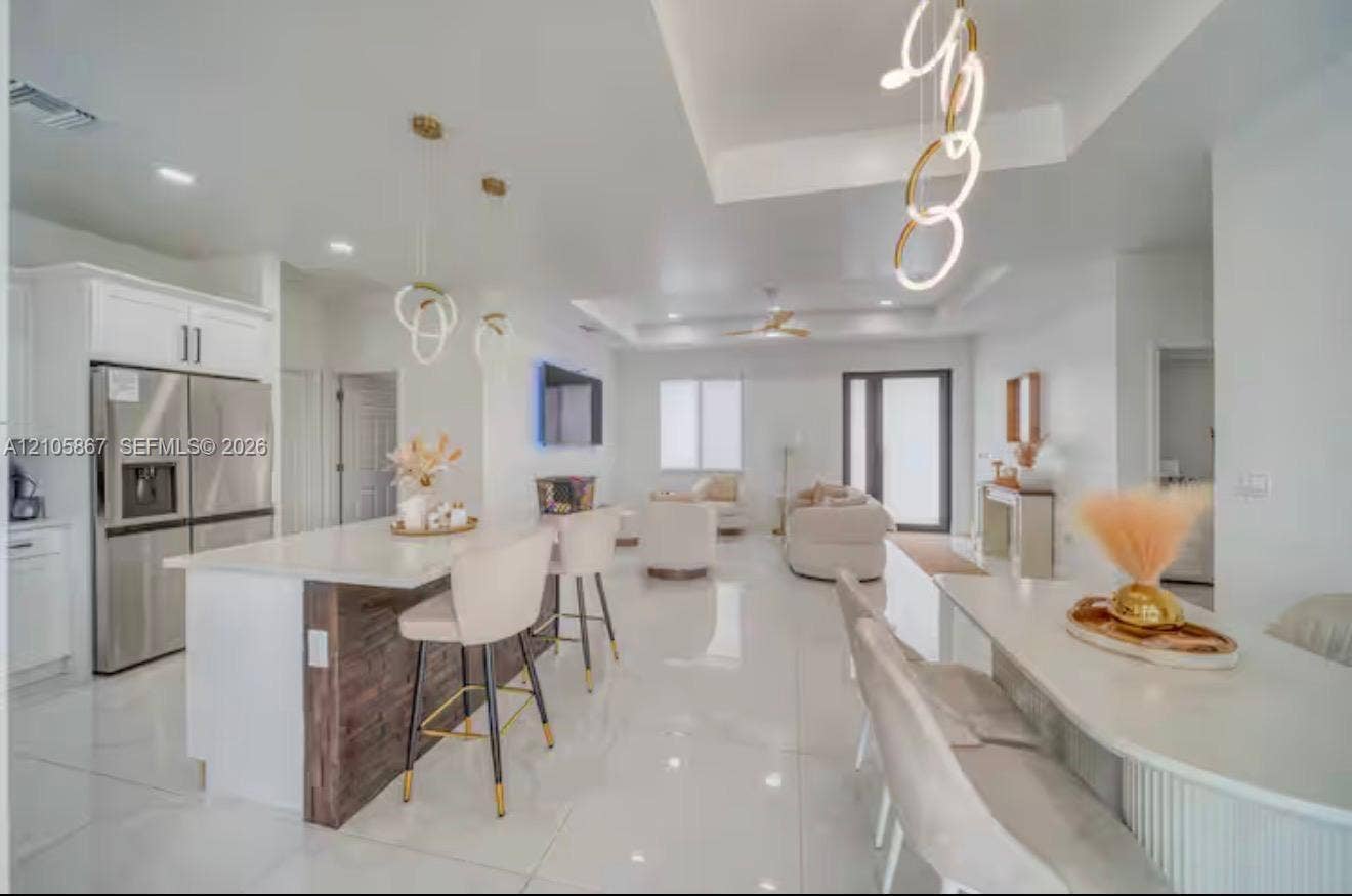 Open-concept living space featuring high-gloss tile flooring, white shaker-style cabinetry, a kitchen island with stone-finish countertops and wood-accented base, tray ceilings, and contemporary ring-style pendant lighting