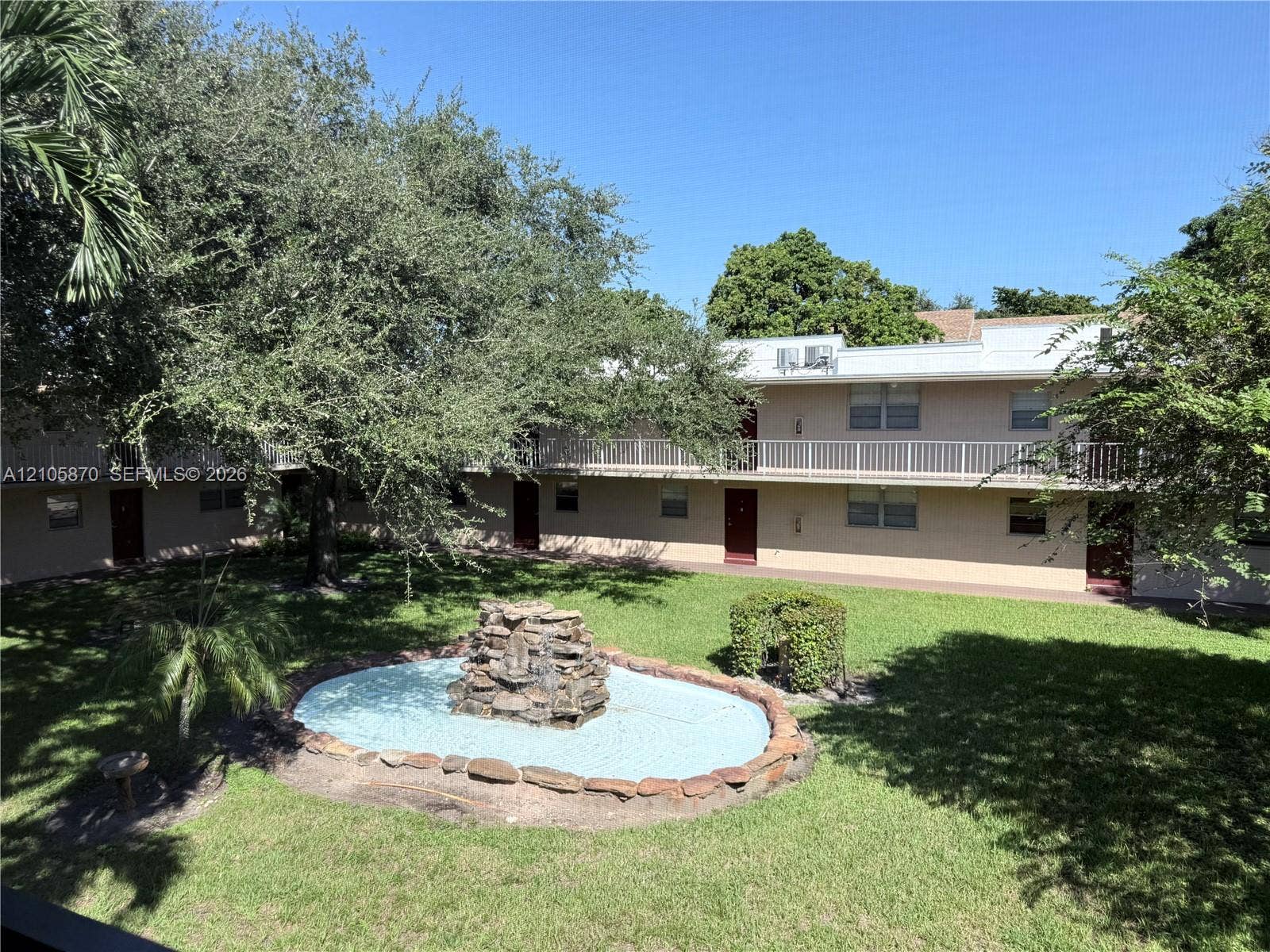 7173 Orange Drive, #204B, Davie, FL 33314 Courtyard featuring a stone-accented fountain, manicured lawn, mature trees, and multi-level exterior walkways with railings