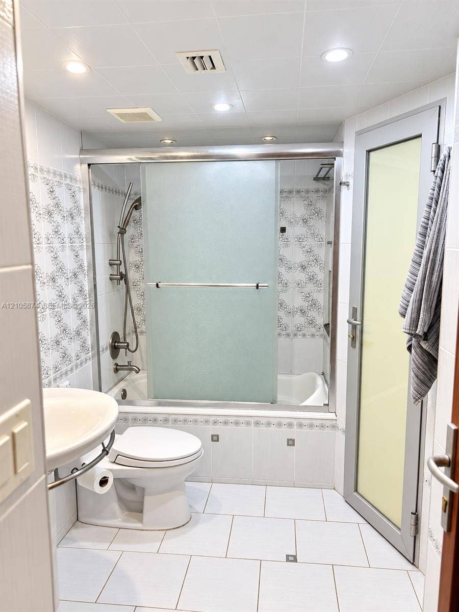 Full-bath featuring a bathtub with a sliding frosted-glass enclosure, wall-mounted shower hardware, decorative floral tile backsplash, ceramic tile flooring, and a pedestal sink